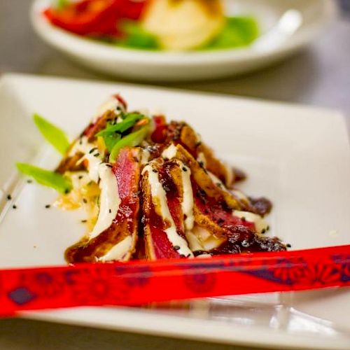 A dish on a white square plate garnished with green onions and red chopsticks beside it. Another plate is blurred in the background.