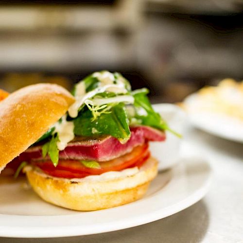 A gourmet burger with fresh vegetables and greens, served with sweet potato fries on a white plate. Another plate of fries is in the background.