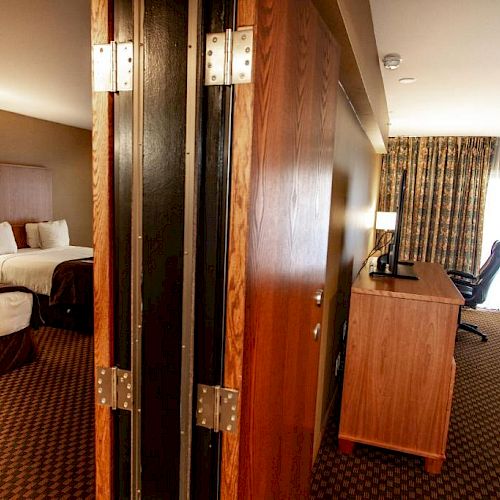 A hotel room showing two areas with beds, separated by a large, partially open wooden door. Each area has furniture and curtains covering windows.