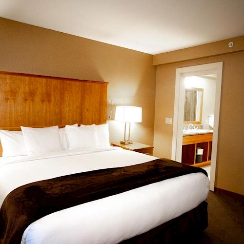 The image shows a hotel room with a large bed, two bedside tables, lamps, and an attached bathroom visible through an open door.