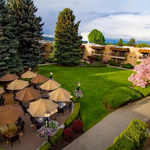 Aerial view of a garden with patio tables and umbrellas surrounded by trees and a walkway, adjacent to a building with balconies.