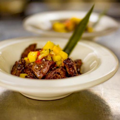 The image shows a bowl of cooked meat pieces topped with diced mango and a green garnish, presented on a white plate, with another bowl blurred in the background.