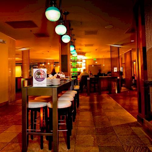 The image depicts a modern bar with high tables and stools, illuminated by hanging lights, and featuring a warm, ambient atmosphere with brown decor.