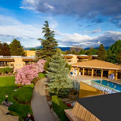 The image shows a scenic view of a resort with lush greenery, a well-maintained garden, umbrellas, a blooming tree, and an outdoor pool area.